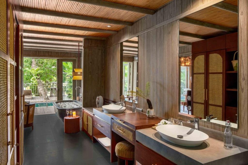 A long timber bathroom with twin white basins and a dressing stool, rattan wardrobes on the left, and a dark stone bathtub with floating petals by doors open onto the pool deck