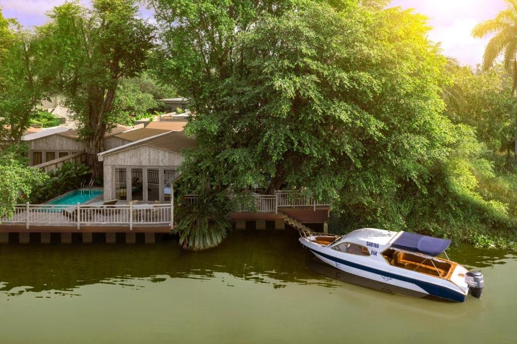 Timber villas with a pool and white railings on a deck over the Saigon River, shaded by a large tree, with steps down to the water and a speedboat moored alongside