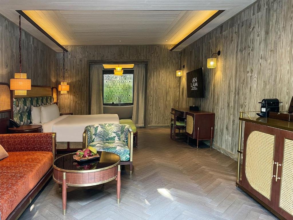 The suite seen from the sofa: a round wooden coffee table with dragon fruit, a leaf-print armchair, the bed and a desk under a wall-mounted TV, and a coffee machine on a rattan cabinet, on a herringbone wood floor