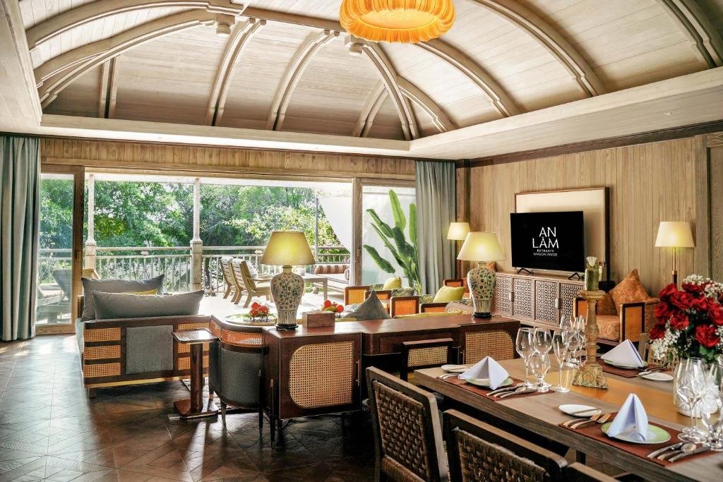 The Signature River View Suite at An Lam Retreats Saigon River: a vaulted timber ceiling over a sofa and armchairs, a TV cabinet with carved doors, a dining table laid with napkins and red roses, and glass doors open onto a balcony among trees