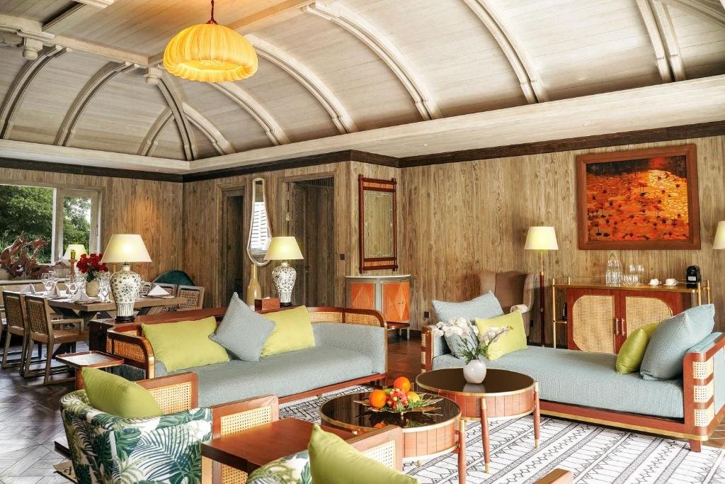 A lounge under a ribbed vaulted ceiling with a silk pendant lamp: two sage daybeds with lime and grey cushions, leaf-print armchairs, round coffee tables with fruit and orchids, and an orange painting on the timber wall