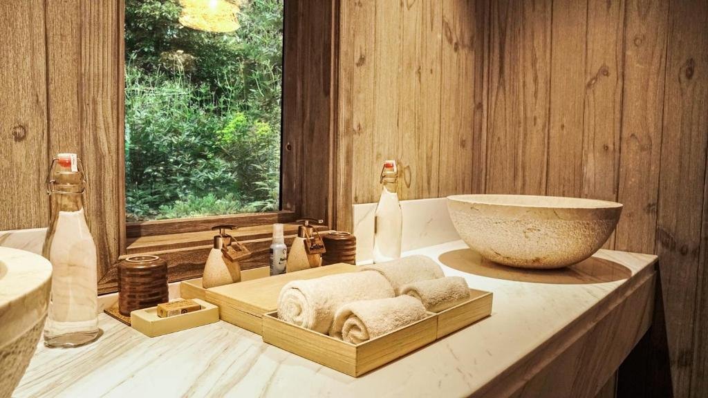 A stone bowl basin on a marble counter with rolled towels, glass bottles and toiletries, beside a window onto green garden