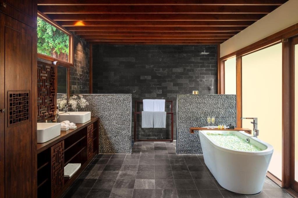 A dark stone bathroom under a timber-beamed ceiling: two white basins on a carved wooden vanity, pebble-stone walls around a walk-in shower, and a freestanding bathtub filled with flower petals and two glasses of champagne