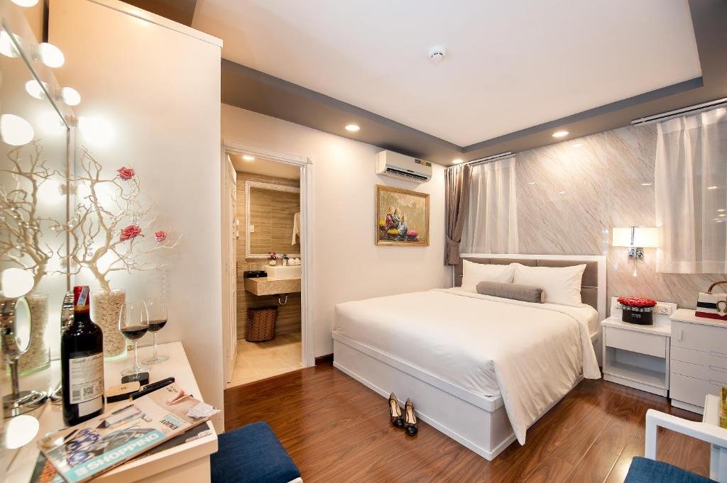 The Deluxe City View Room at Adora Hotel: a double bed with a grey bolster against a marble-effect wall with sheer curtains, a white vanity with bulb lights and a bottle of wine in front, and the bathroom through an open door