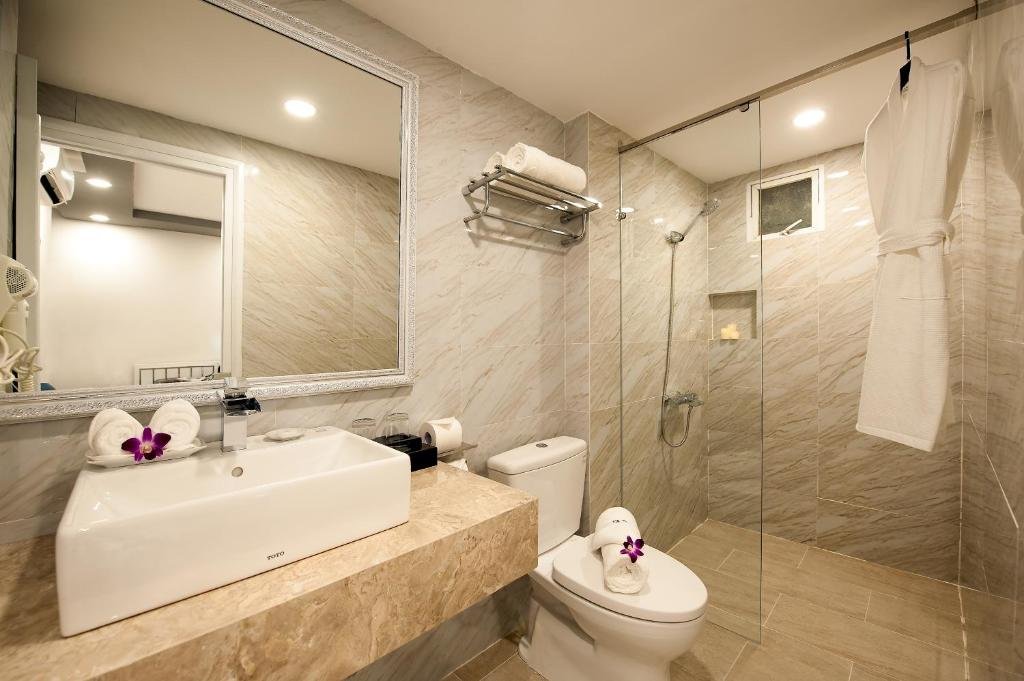 A marble-tiled bathroom with a basin on a stone counter under a large mirror, rolled towels with orchids, a toilet, a bathrobe on a hook, and a glass walk-in shower