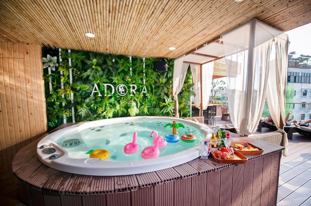 A round hot tub with inflatable flamingo drink holders and a tray of fruit and drinks, under a bamboo ceiling in front of a green plant wall with the Adora sign, and curtained daybeds on the roof terrace beyond