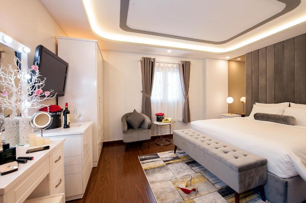 The Premium King Room at Adora Hotel: a king bed against a tall grey velvet headboard with a tufted bench at its foot, a grey armchair by a curtained window, and a white vanity with bulb lights, red roses and a bottle of wine