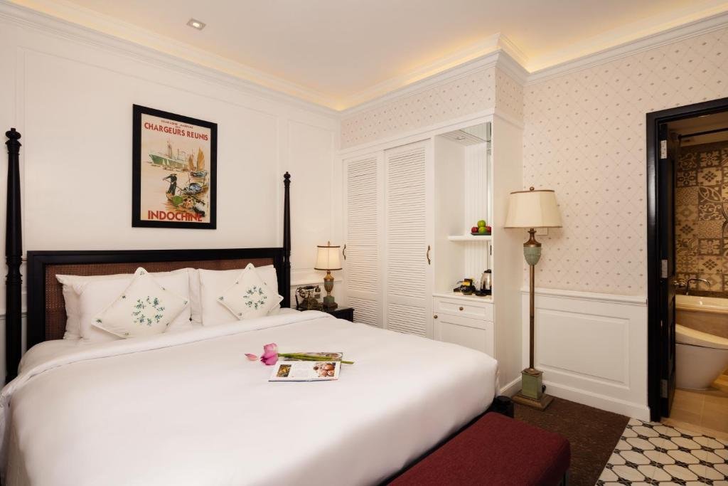 The four-poster bed with embroidered cushions under a Chargeurs R&eacute;unis Indochine poster, white louvred wardrobes with a kettle and fruit, a standing lamp, and the bathroom through a black door