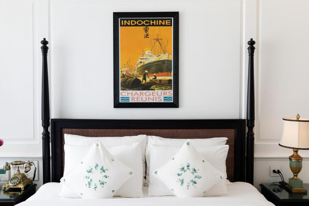 A vintage Indochine shipping poster above the black four-poster headboard with rattan panels, embroidered cushions, a brass antique-style telephone and a green-and-gold lamp