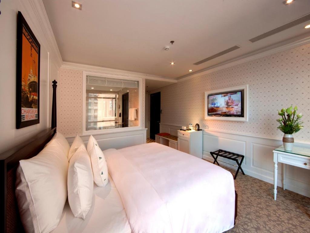 The king bed in the foreground, a glass panel into the bathroom, a TV in a white frame, a kettle cabinet, a luggage rack and a small desk with lotus flowers on a patterned carpet