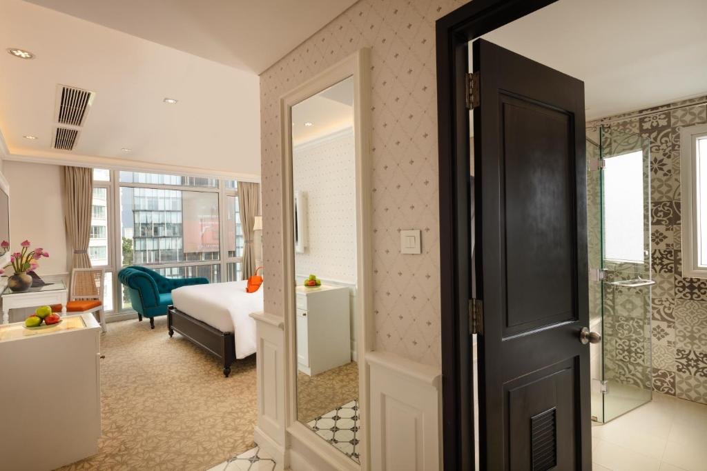 The suite seen from the entrance past a tall mirror: the bed, teal chaise and windows beyond, and the bathroom with its glass shower through a black door on the right