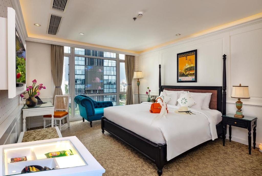 The Imperial Heritage Suite at Alagon D&rsquo;antique: a dark four-poster king bed under an Indochine shipping poster, a teal tufted chaise before floor-to-ceiling windows onto city towers, a white desk with lotus flowers, and a TV on the wall
