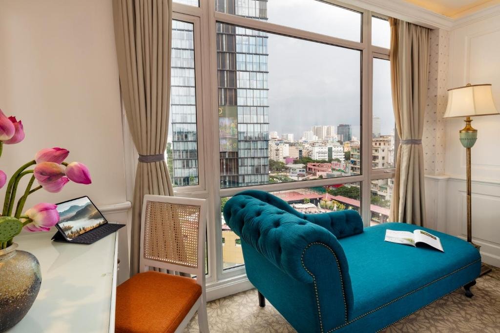 A teal velvet chaise longue at a tall corner window over District 1, a white desk with a rattan chair, a tablet and pink lotus flowers, and a green-and-gold standing lamp
