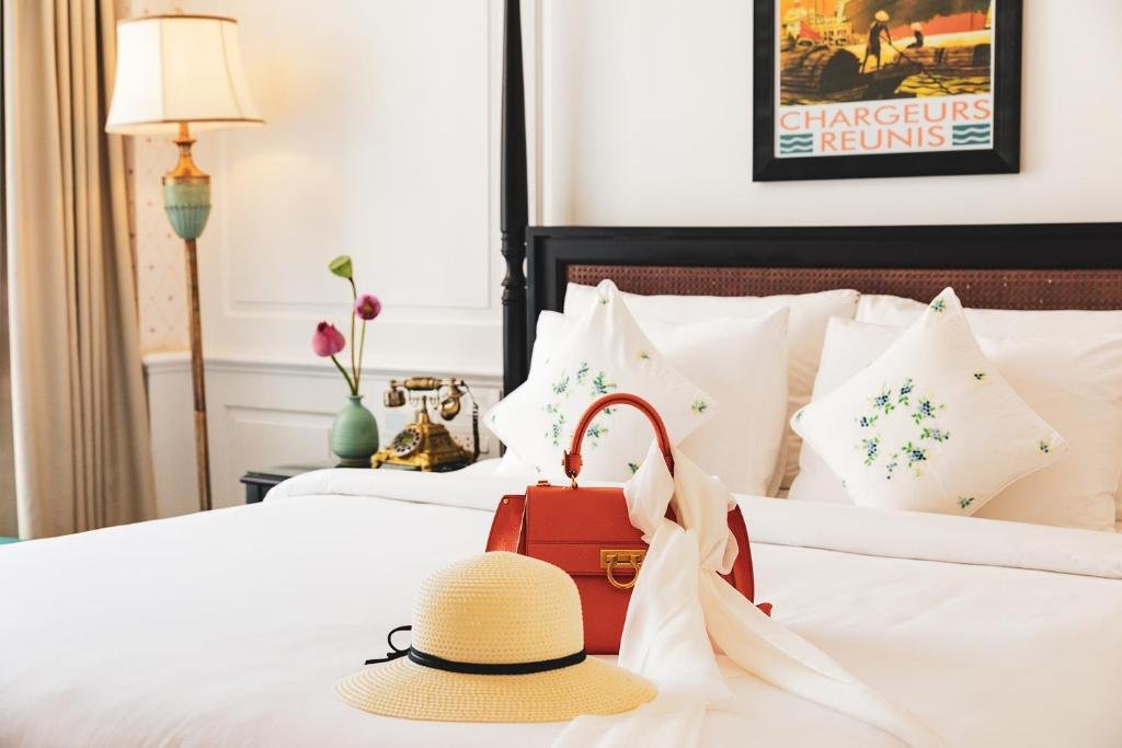 A straw hat and a red handbag on the white bed with embroidered cushions, a brass antique-style telephone and lotus flowers on the bedside table, and a poster above