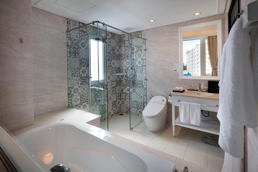 A bathroom with a bathtub in the foreground, a glass walk-in shower lined with patterned tiles and a small window, a smart toilet, and a white vanity under a mirror with a bathrobe hanging nearby