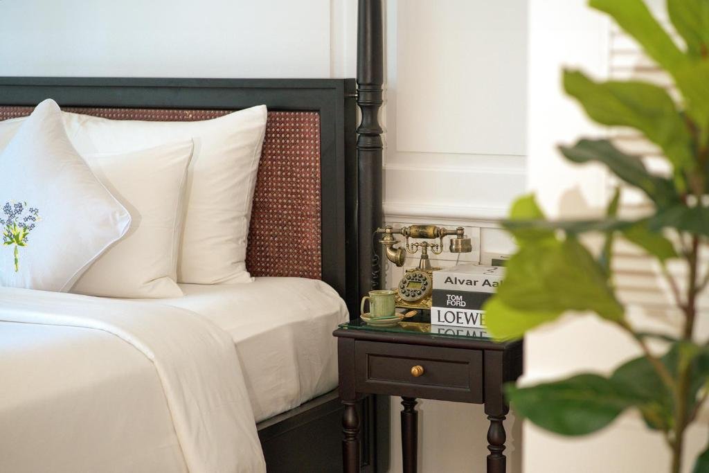 The rattan-panelled headboard with an embroidered cushion, a brass antique-style telephone, a green cup and design books on the dark bedside table, and a fiddle-leaf fig in the foreground