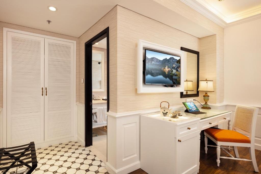 White louvred wardrobe doors, the bathroom through a black-framed doorway, a TV in a white frame above a desk with a tea set and a tablet, and patterned cement tiles by the entrance