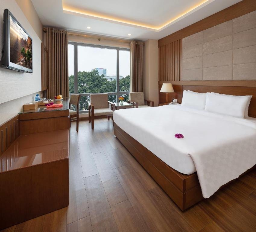 The Premium Deluxe Double at EdenStar Saigon: a large bed against a padded headboard and wood-slatted wall, a long wooden desk and TV, and three armchairs at a floor-to-ceiling window onto treetops and the city