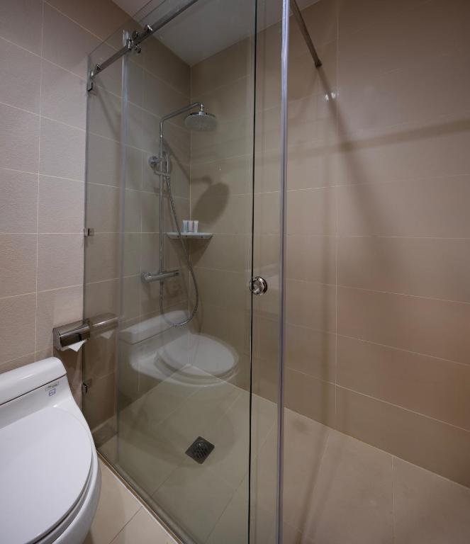 A glass-screened shower with a rain shower head and hand shower on beige tiles, beside the toilet