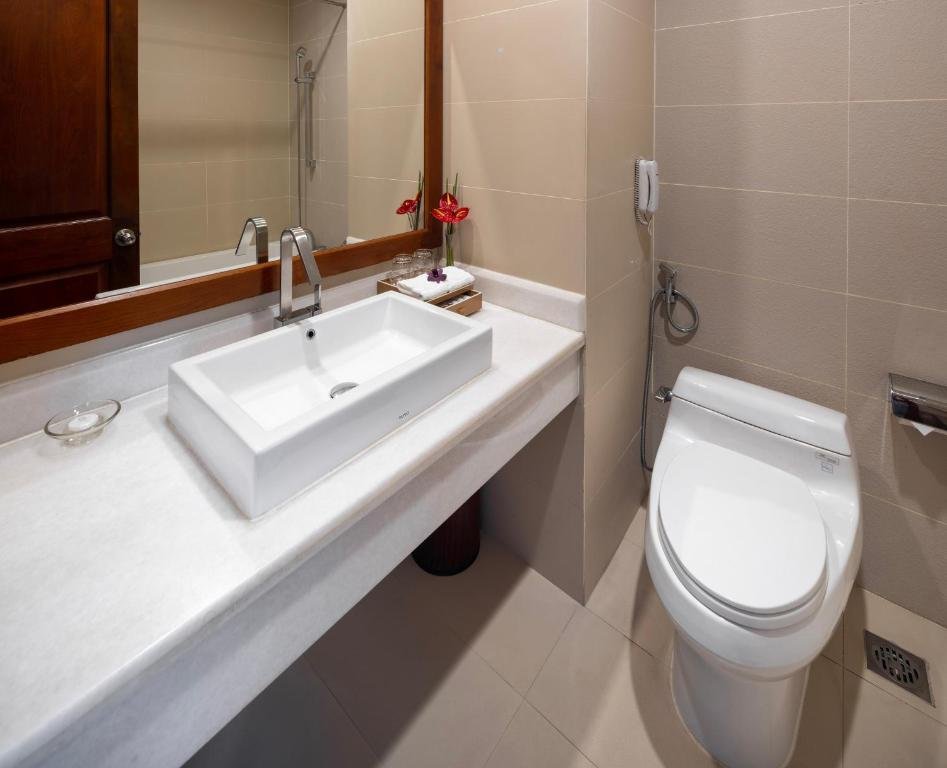 A white rectangular basin on a marble counter under a wood-framed mirror with red anthurium flowers and folded towels, a toilet and a wall hairdryer