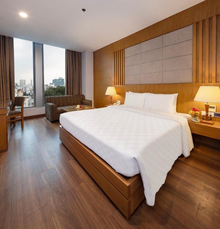 The Executive Double at EdenStar Saigon: a large bed against a padded panel on a wood-slatted wall, bedside lamps and flowers, and a striped sofa and coffee table by two tall windows onto the city