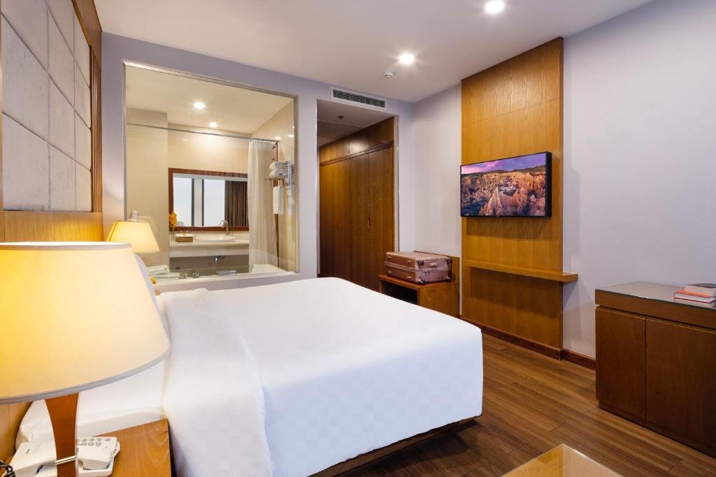 The bed with a bedside lamp and telephone, a glass wall into the bathroom with its bathtub and basin, a wall-mounted TV on an oak panel, and a suitcase stand by the wardrobe