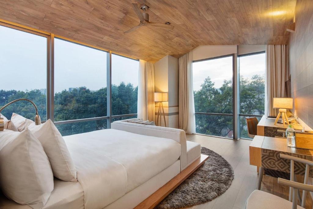 The bed with a white sofa at its foot on a round shaggy rug, a tripod lamp and a desk, and glass on two sides looking onto trees
