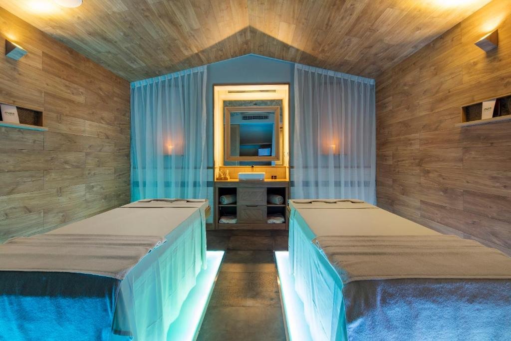 A spa room for two with lit massage beds, sheer curtains, a small basin and rolled towels under a pitched timber ceiling