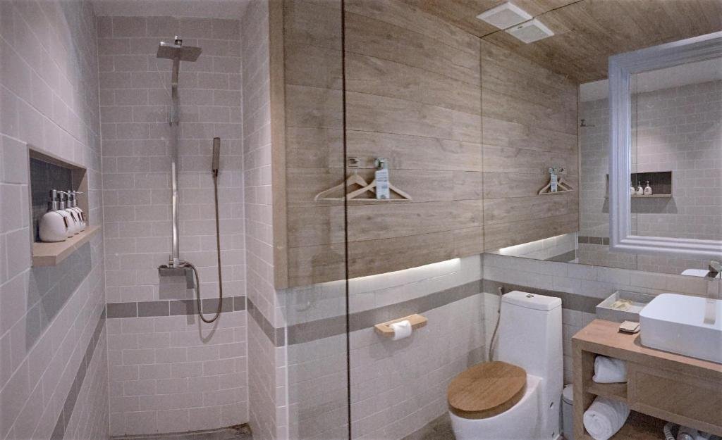 A bathroom with white subway tiles: a walk-in rain shower with a niche of toiletries behind glass, a toilet with a wooden seat, and a basin on a wooden stand under a white-framed mirror