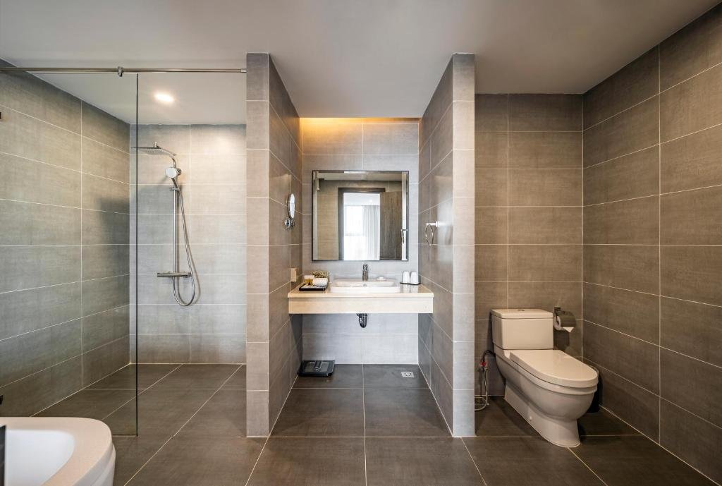 The bathroom in grey stone tiles: a glass-screened walk-in rain shower, a basin under a lit mirror, and a toilet in its own bay