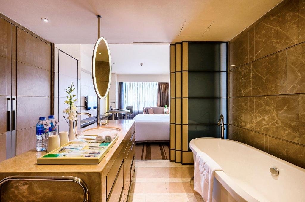 A long marble vanity with a round lit mirror and a tray of amenities, a freestanding bathtub on the right, and the bed and window seen through the open doorway