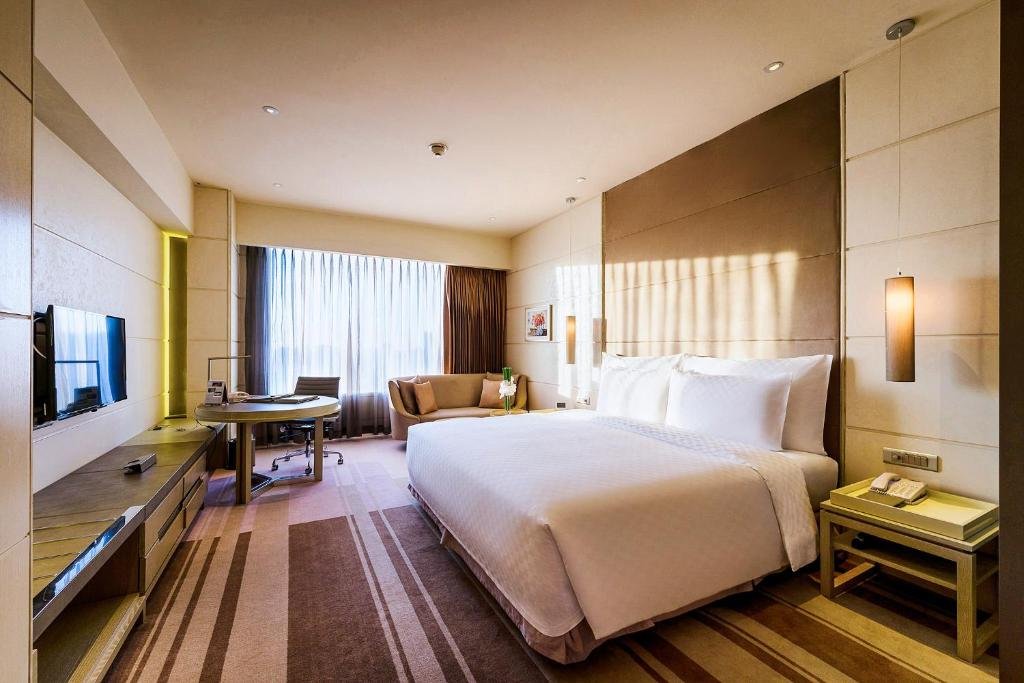 The Deluxe Premium King room at Hotel Nikko Saigon: a king bed against a tall padded headboard, a round desk with an office chair, a sofa under a wide curtained window, and a TV on a long low console