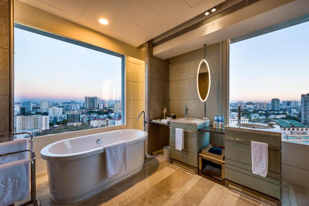 A marble bathroom with a freestanding bathtub under a picture window over the Saigon skyline at dusk, two basins on grey-green vanities, a round lit mirror and a towel rail
