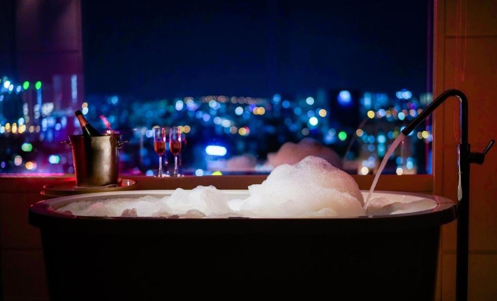 A freestanding bathtub full of bubbles by a window at night, with the city lights blurred beyond, a champagne bucket and two glasses of rosé on the sill