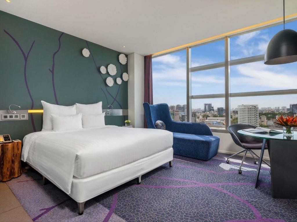 The Superior King Room at Pullman Saigon Centre: a king bed against a teal wall with a painted branch and white paper flowers, a blue wingback chaise, and a round glass desk by floor-to-ceiling windows over the city