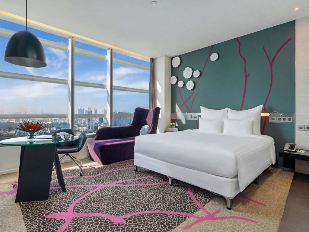 The Deluxe King Room at Pullman Saigon Centre: a king bed against a teal wall with a magenta painted branch and white paper flowers, a purple chaise and a round glass desk at a corner of floor-to-ceiling windows over the city