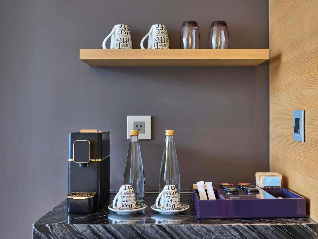An espresso machine, two espresso cups, glass water bottles and a tray of coffee pods on a marble counter, with mugs and glasses on a shelf above