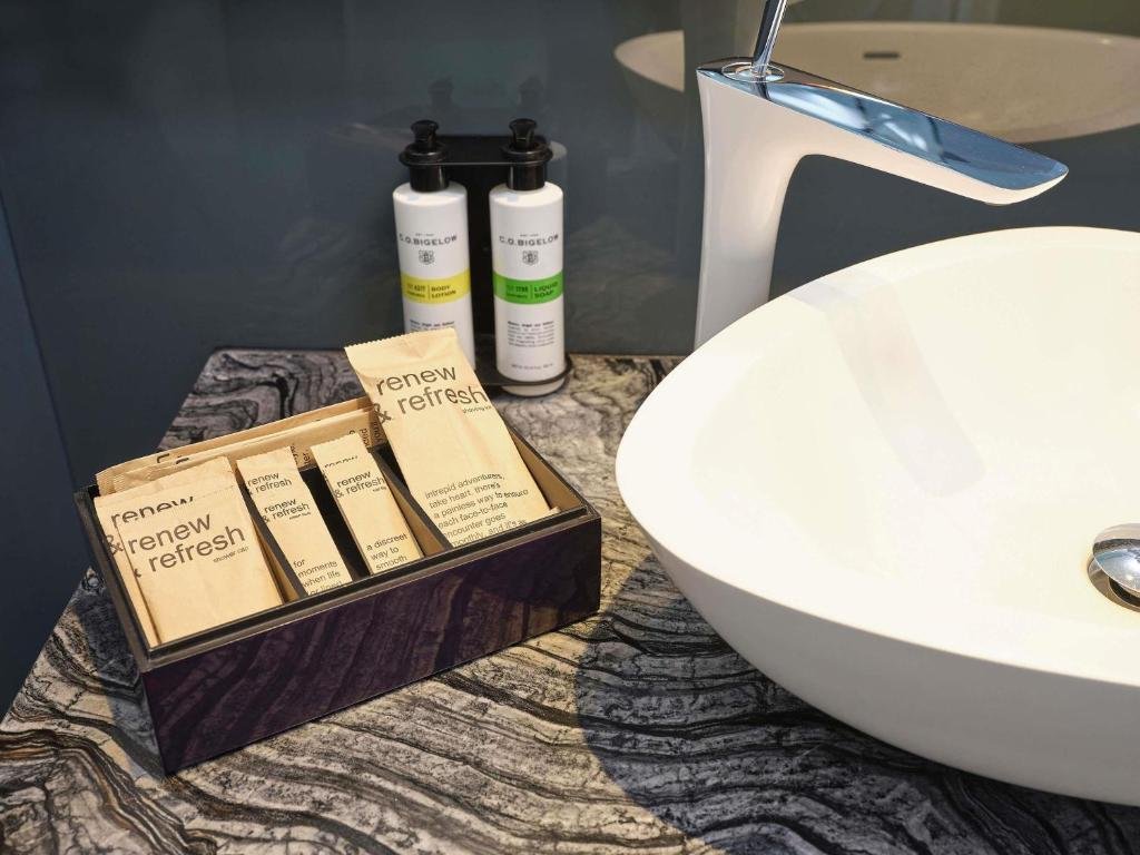 C.O. Bigelow body lotion and liquid soap beside a box of amenity sachets on a marble counter, next to a white bowl basin and a curved tap