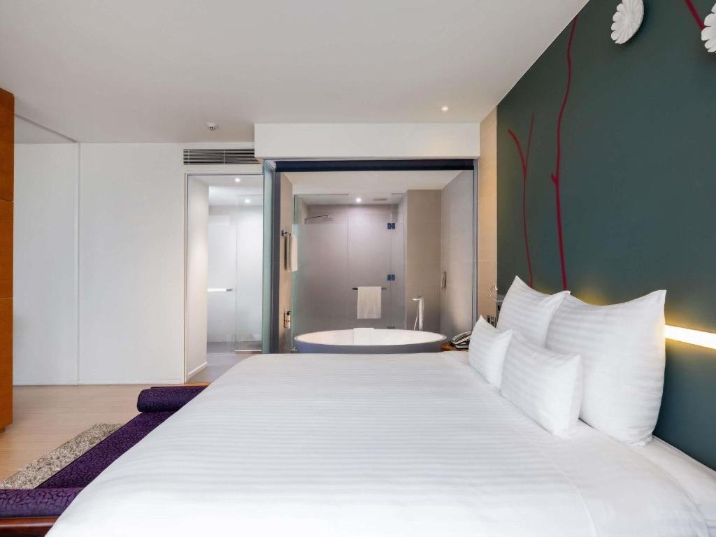 A king bed with a purple bench at its foot against a teal wall with a red painted branch, and through a glass wall a round white bathtub and a glass shower