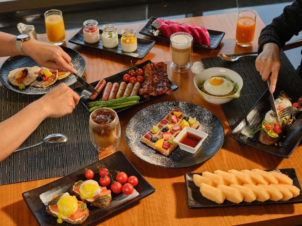 Two guests at a breakfast spread of eggs Benedict, avocado toast, a fruit waffle, pancakes, sausages, bacon, dragon fruit, melon and orange juice