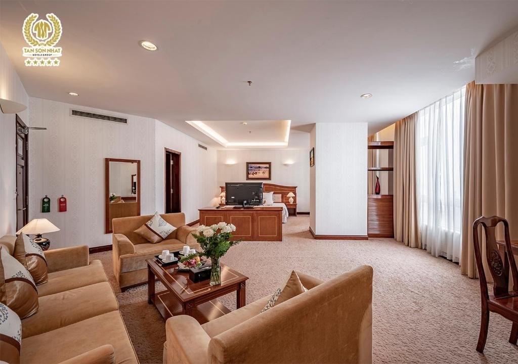 The Suite at Tan Son Nhat Saigon Hotel: a living room with beige sofas around a wooden coffee table set with white roses and a tea service, and the bed and TV cabinet at the far end under a lit ceiling cove