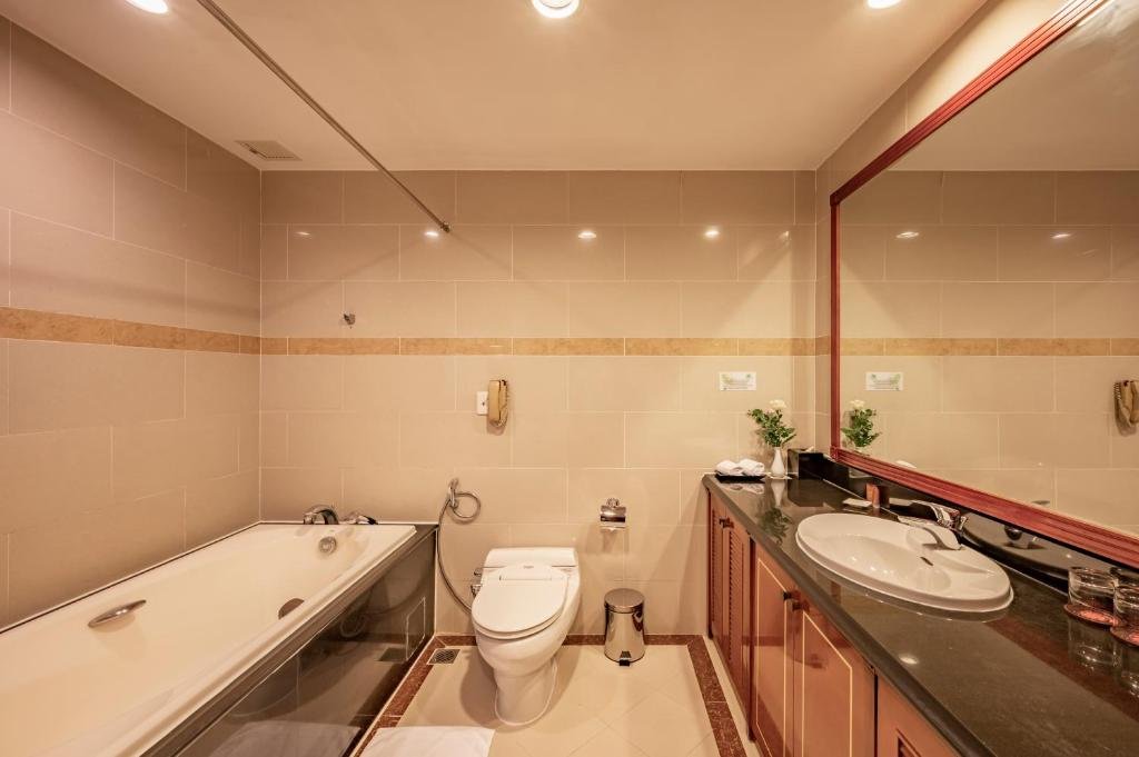 The Suite bathroom: a bathtub on the left, a toilet, and a long dark stone vanity with a basin under a wood-framed mirror, on beige tiles with a patterned border