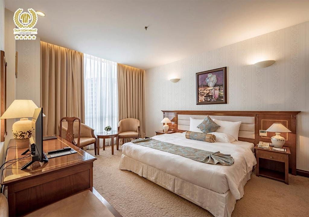 The Suite Deluxe bedroom: a king bed with a patterned runner and bolster under a framed painting, bedside lamps on wooden tables, and two armchairs by a tall curtained window
