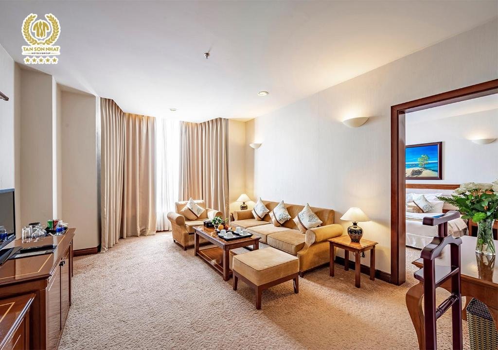 The Suite Deluxe living room: a beige sofa and armchair around a wooden coffee table with a fruit plate, a TV cabinet on the left, and the bedroom visible through a wooden doorway on the right