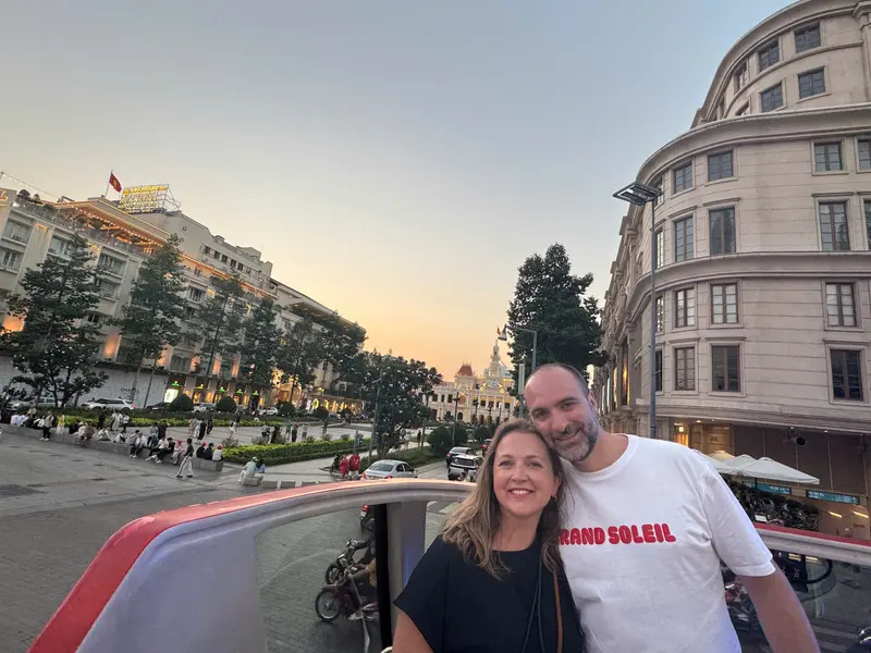 Two guests on the open top deck of a Saigon sightseeing bus at dusk, with a hotel and its rooftop sign on the left, the colonial City Hall at the end of Nguyen Hue boulevard and a curved stone building on the right