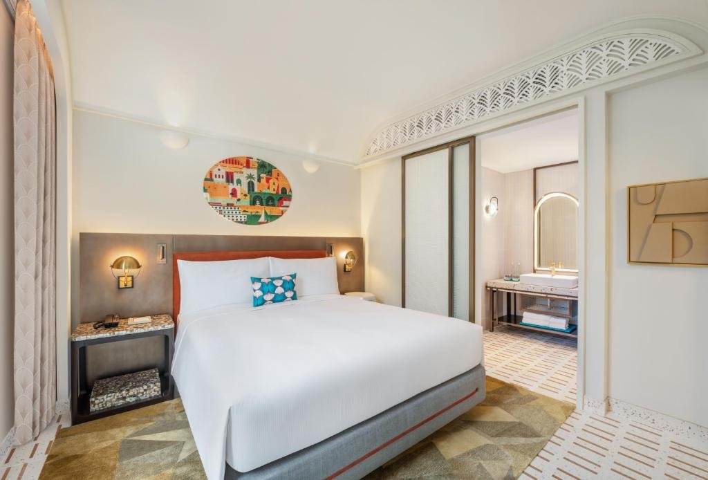 Deluxe King Room at La Festa: a king bed between two brass wall lights under a round canvas of a Mediterranean harbour village, with glazed doors under a fretwork arch opening to the bathroom vanity