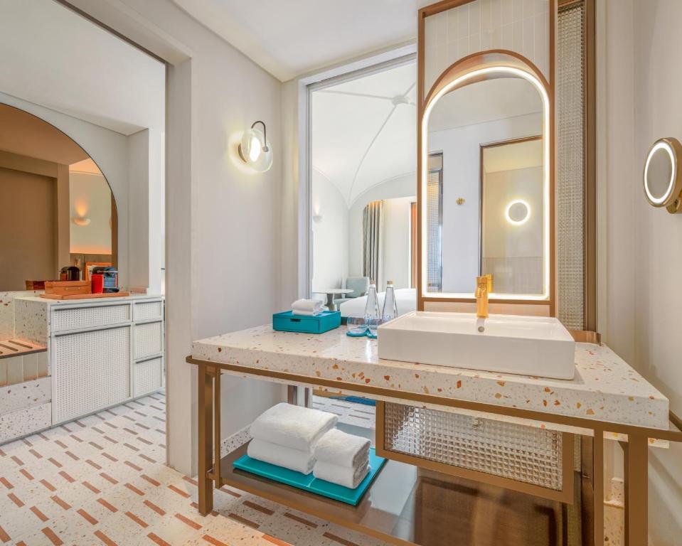 The Classico Ocean View bathroom: a terrazzo counter on a brass frame with a square basin and gold tap, a backlit arched mirror above and folded towels on the shelf below, with the bedroom visible through the doorway