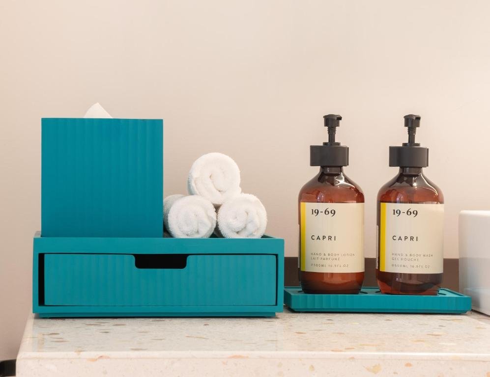 Rolled white towels in a teal box beside 19-69 Capri hand lotion and body wash on the terrazzo counter