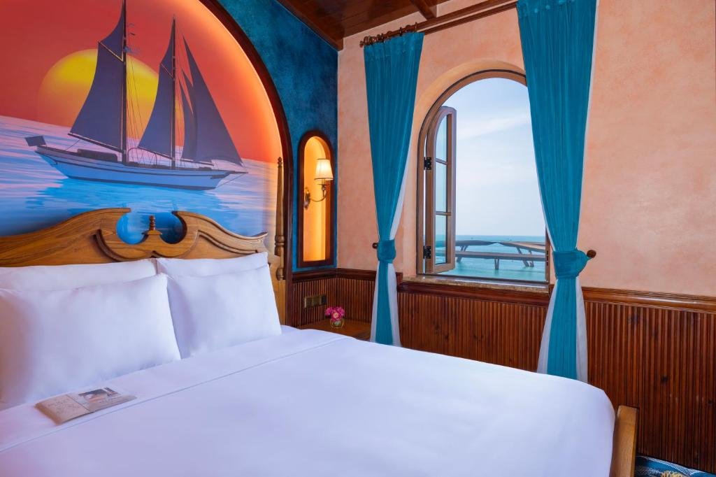 The bed beneath a sunset mural of a sailing ship, with an arched window beside it opened onto the sea and a curving footbridge over the water
