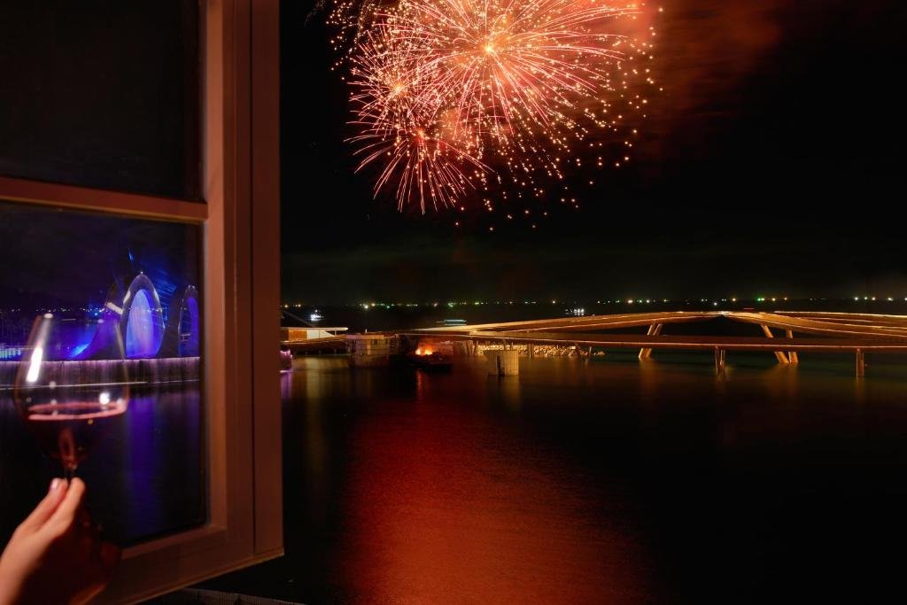 Fireworks bursting over the sea at night, seen past a raised glass of wine at the open window, with the lit curve of a footbridge on the water below